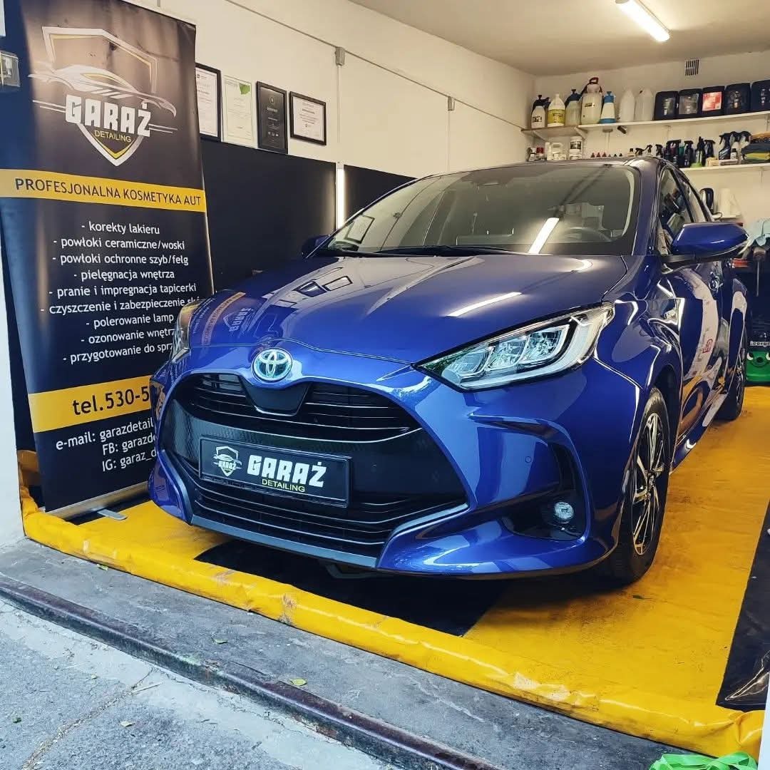 Błyszczący, niebieski Toyota Yaris po detailingu, z widocznym logo firmy 'Garaż Detailing' na banerze i tablicy rejestracyjnej, na żółtym podłożu w dobrze oświetlonym garażu.