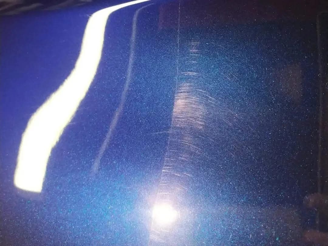 Błyszczący, metaliczny lakier samochodowy w kolorze niebieskim z widocznymi drobnymi zarysowaniami i hologramami po polerowaniu, odbicie światła tworzy białą smugę.