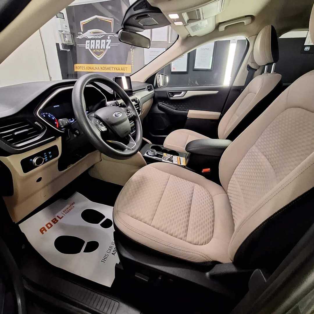 Wnętrze Forda Escape po detailingu, z beżową tapicerką foteli o wzorze rombów, widoczne logo detailingowe na ścianie i papierowa mata podłogowa z nadrukowanymi śladami stóp.