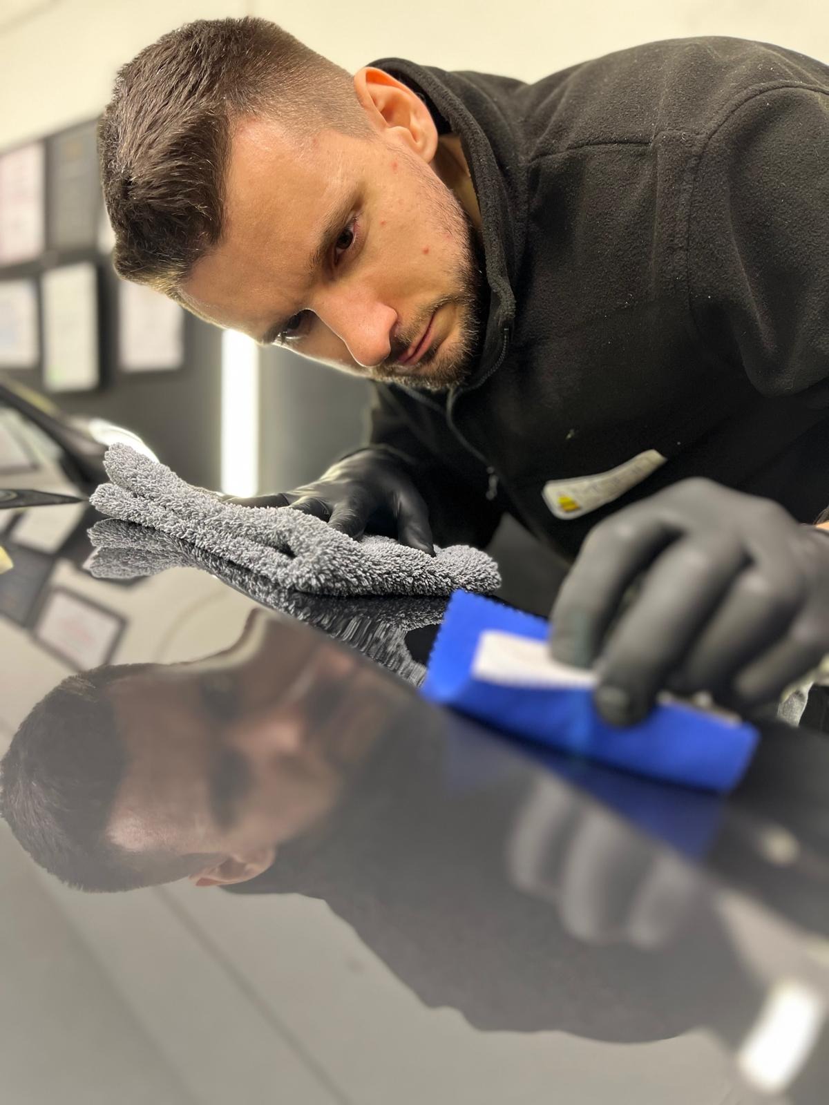 Detailer w czarnych rękawiczkach poleruje lakier ciemnego samochodu przy użyciu niebieskiego aplikatora i szarej mikrofibry, widoczne odbicie twarzy detailera w lakierze.