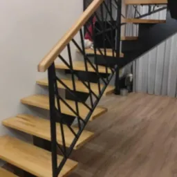 Drewniane schody z czarną, geometryczną balustradą metalową i jasną drewnianą poręczą w nowoczesnym wnętrzu.