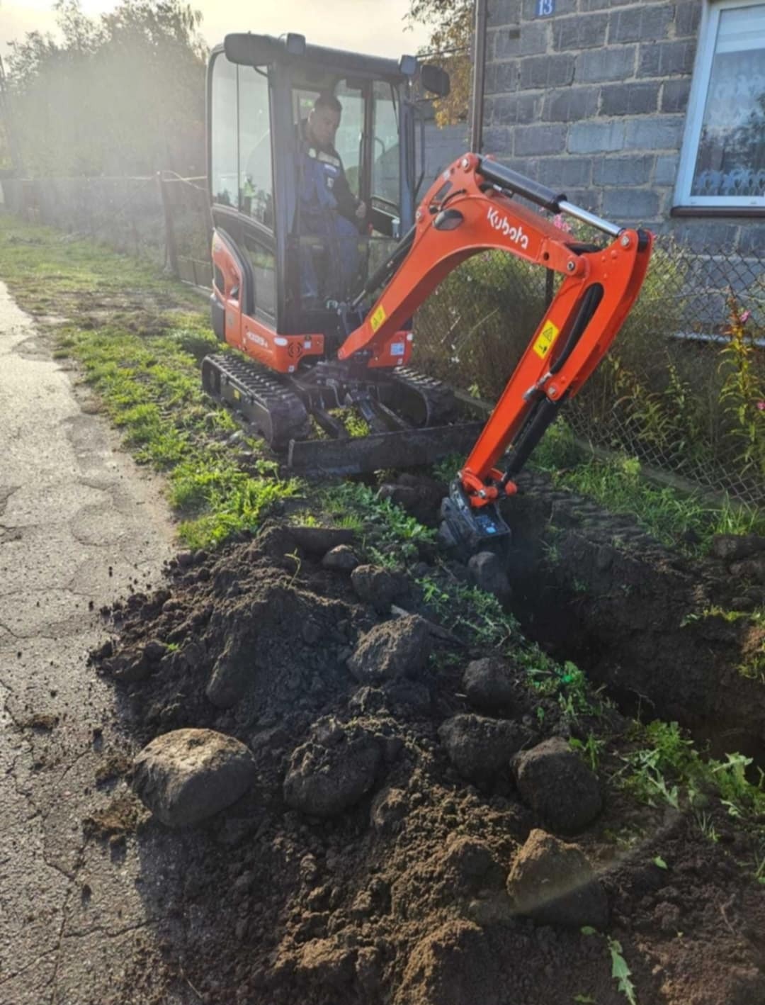 Pomarańczowa mini koparka Kubota z operatorem w trakcie prac ziemnych przy posesji z numerem 13, widoczny wykop i sterta ziemi z kamieniami.