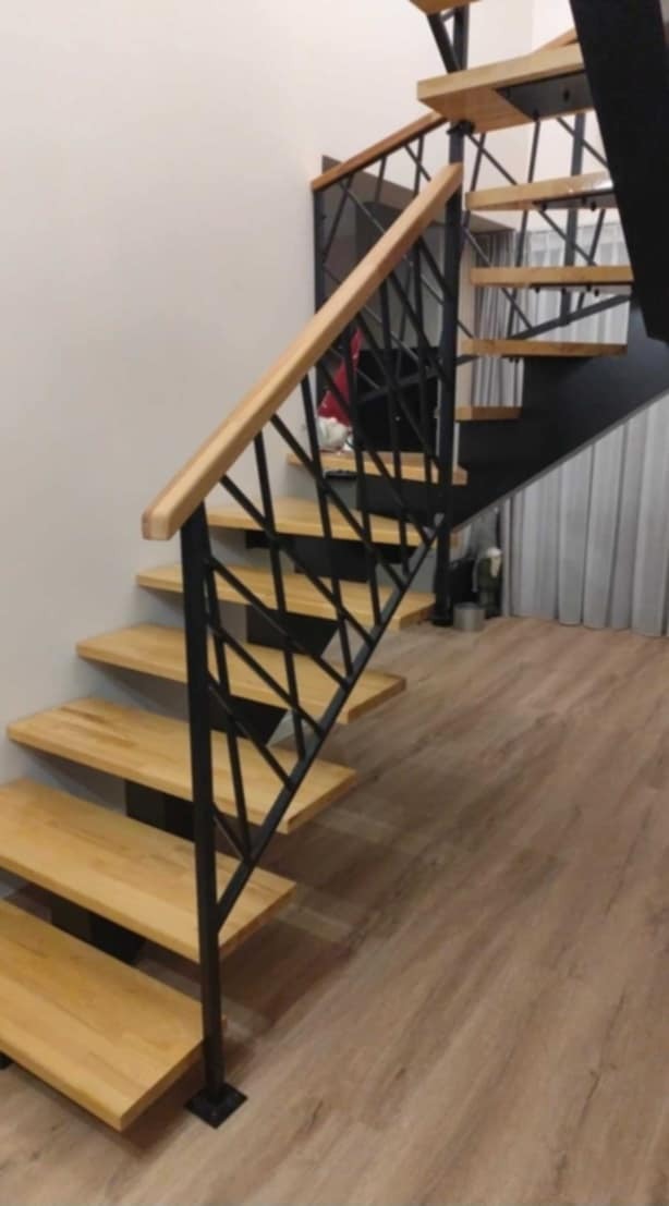 Drewniane schody z czarną, geometryczną balustradą metalową i jasną drewnianą poręczą w nowoczesnym wnętrzu.