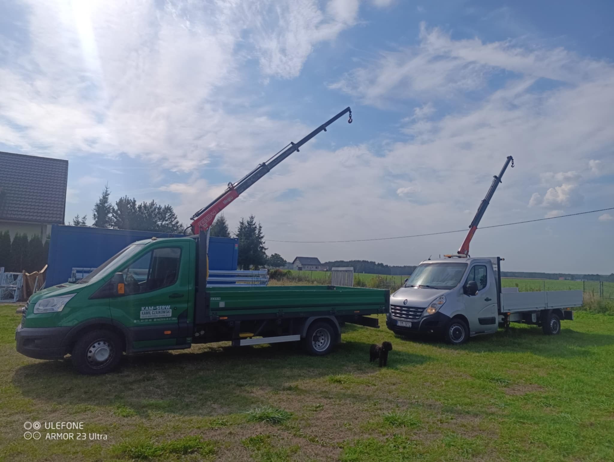 Dwa samochody dostawcze z zamontowanymi dźwigami na trawiastym terenie; jeden zielony z logiem firmy KAM-SERW, drugi srebrny, oba z wysuniętymi ramionami dźwigów; w tle zabudowania i błękitne niebo...