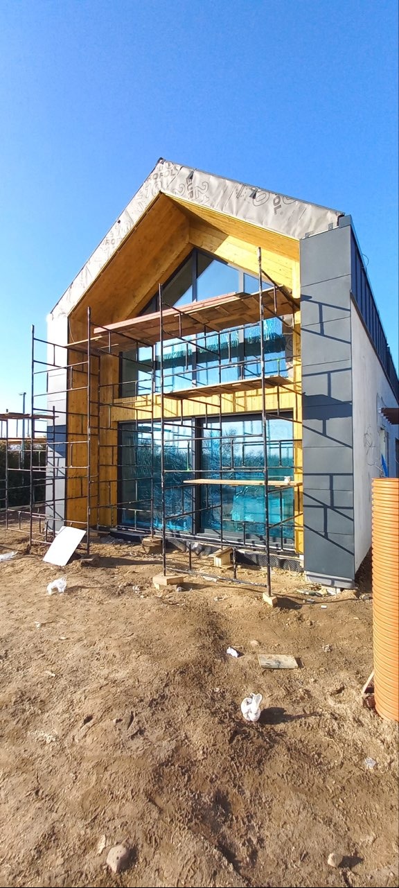 Surowy stan budowy domu z drewnianą konstrukcją szkieletową i zamontowanymi oknami, widoczne rusztowania i teren budowy w trakcie prac.