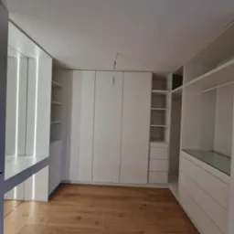 Przestronna garderoba z białymi szafami wnękowymi z ryflowanymi frontami, oświetleniem LED i drewnianą podłogą w trakcie montażu, widoczna żarówka na kablu.