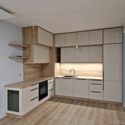 Linea Premium - Nowoczesna kuchnia narożna z frontami w odcieniach beżu i drewna, czarnymi uchwytami i oświetleniem LED pod szafkami wiszącymi, widok na drewnianą podłogę.