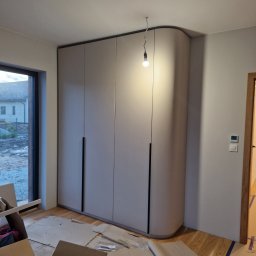 Linea Premium - Montaż szafy wnękowej we Wrocławiu: widok na dużą, beżową szafę z zaokrąglonym bokiem i czarnymi uchwytami, widoczne kartony i materiały zabezpieczające podłogę, surowa żarówka zwisająca z sufitu.