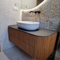 Linea Premium - Nowoczesna łazienka z wiszącą szafką z lamelami, umywalką nablatową i podświetlanym lustrem w kształcie zaokrąglonego prostokąta na tle dekoracyjnych płytek.