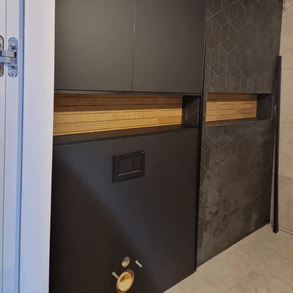 Czarna zabudowa toalety podtynkowej z drewnianymi akcentami i geometryczną płytką w tle. Widoczny przycisk spłukiwania i elementy instalacji. Drzwi częściowo otwarte po lewej stronie.