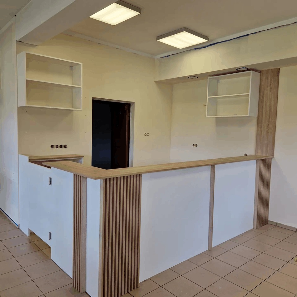 Nowoczesna, częściowo ukończona zabudowa kuchenna z białymi szafkami i drewnianymi panelami dekoracyjnymi, widoczne niedokończone wykończenia i brak sprzętów AGD.