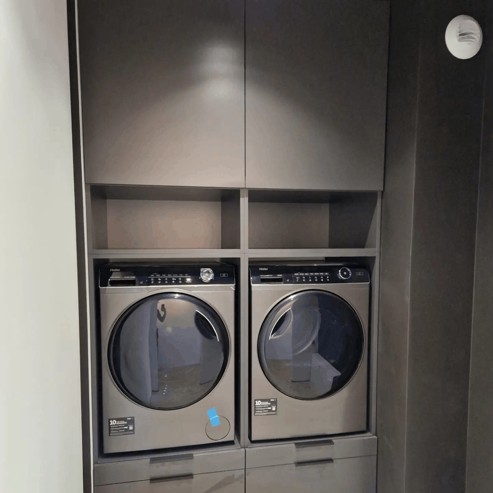 Szara zabudowa meblowa z pralką i suszarką Haier, górne szafki zamykane, dolne z szufladami, oświetlenie sufitowe, nowoczesny design.