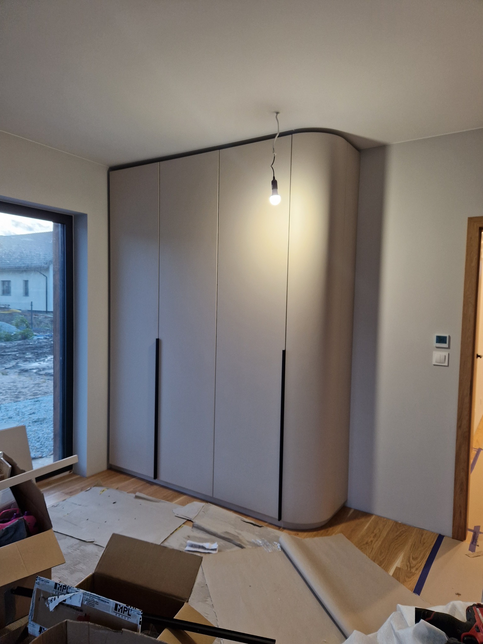 Montaż szafy wnękowej we Wrocławiu: widok na dużą, beżową szafę z zaokrąglonym bokiem i czarnymi uchwytami, widoczne kartony i materiały zabezpieczające podłogę, surowa żarówka zwisająca z sufitu.