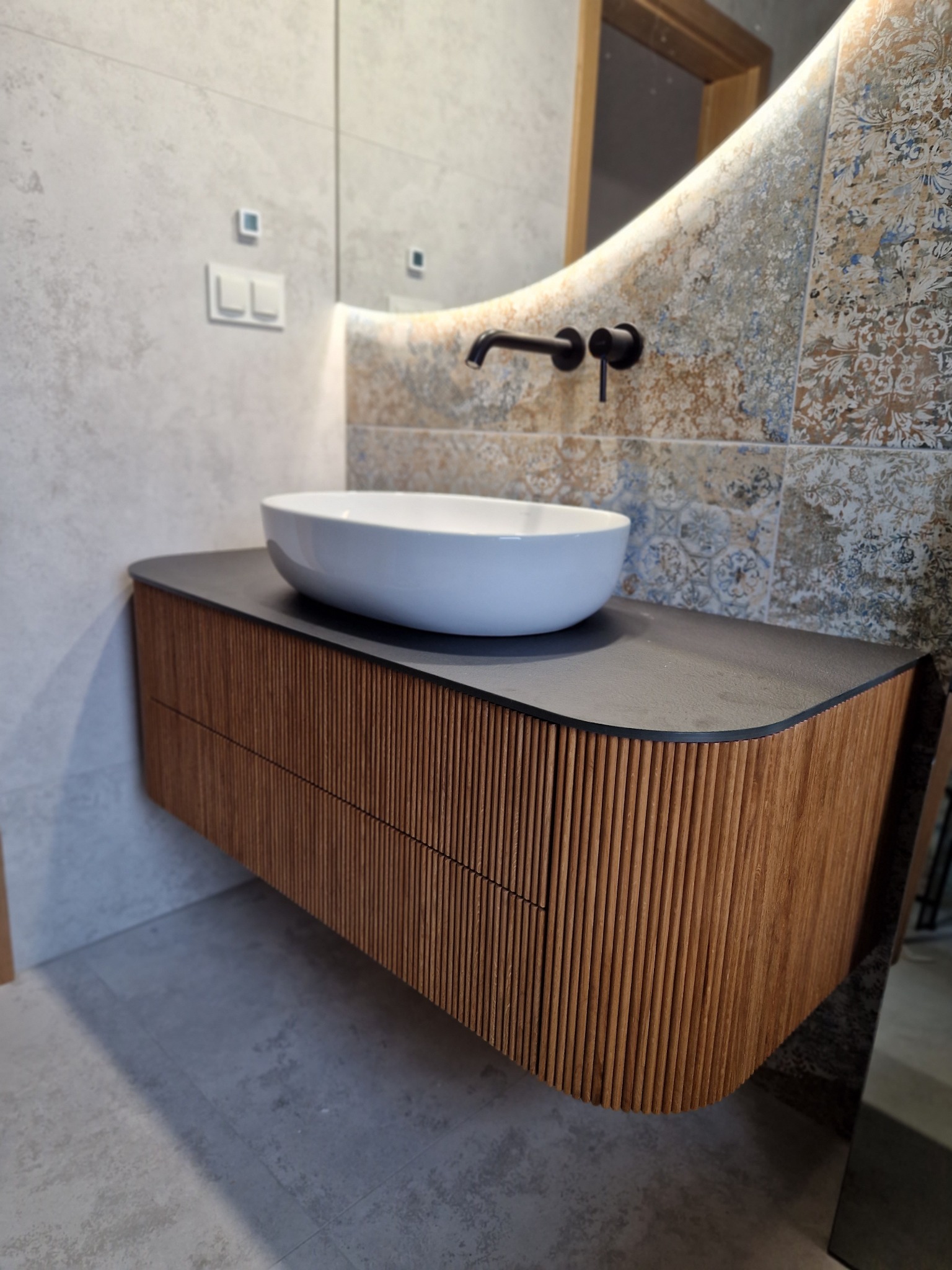 Nowoczesna łazienka z wiszącą szafką z lamelami, umywalką nablatową i podświetlanym lustrem w kształcie zaokrąglonego prostokąta na tle dekoracyjnych płytek.