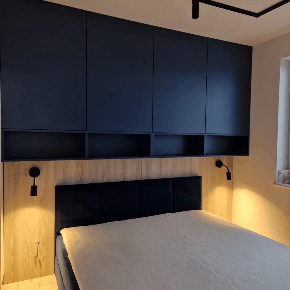 Nowoczesna sypialnia z zabudową meblową: granatowe szafki wiszące z otwartymi półkami nad wezgłowiem łóżka, drewniana boazeria, czarne kinkiety ścienne, biała pościel z pikowanym wzorem.