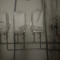 M-BUD Marcel Żak - Trzy nowe grzejniki, jeszcze w folii ochronnej, zamontowane na ścianie z widocznymi rurami instalacji centralnego ogrzewania.