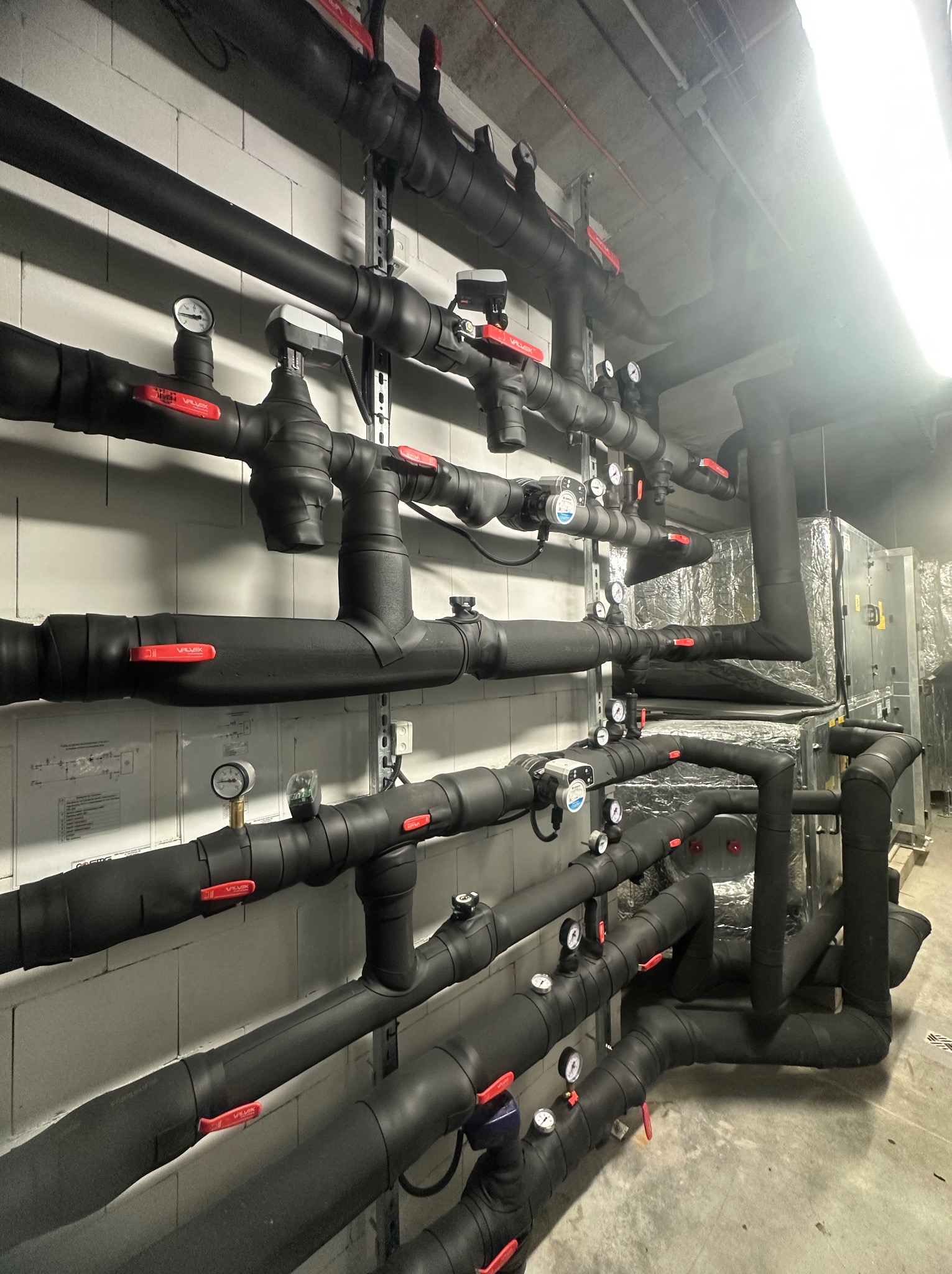 Czarno izolowane rury z czerwonymi zaworami i manometrami zamontowane na ścianie z szarej cegły, widoczna instalacja wentylacyjna w tle.