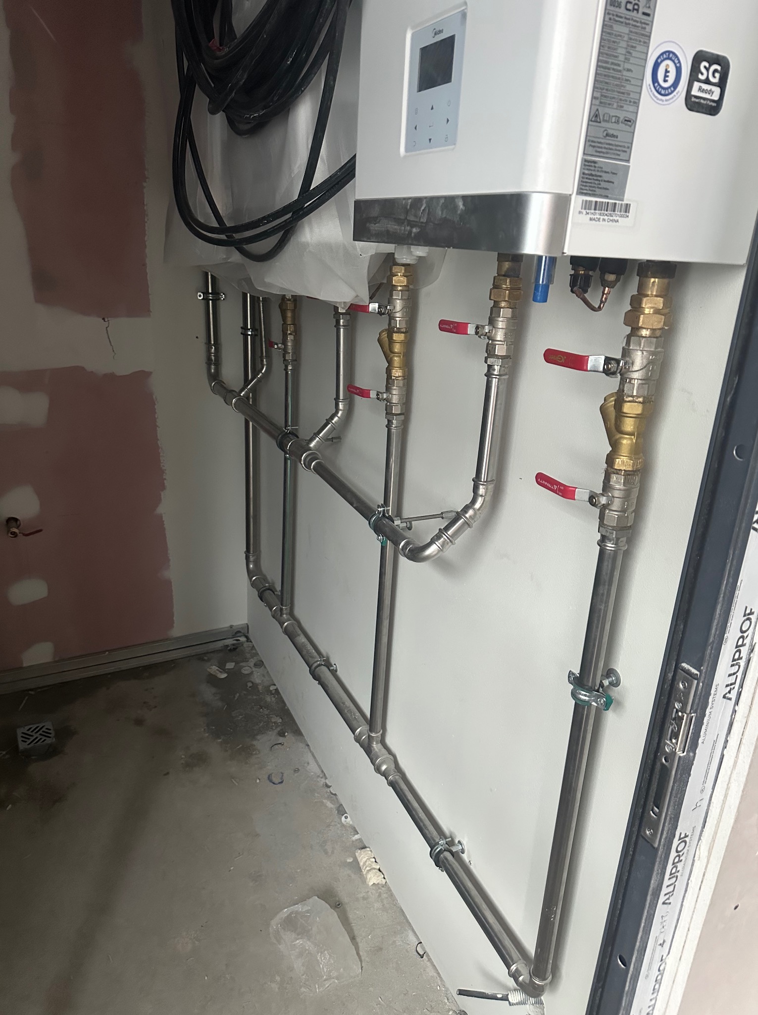 Nowa instalacja grzewcza z rurami ze stali nierdzewnej, zaworami z czerwonymi uchwytami i białym piecem Midea zamontowana na ścianie w surowym pomieszczeniu z niepomalowanymi ścianami i betonową...