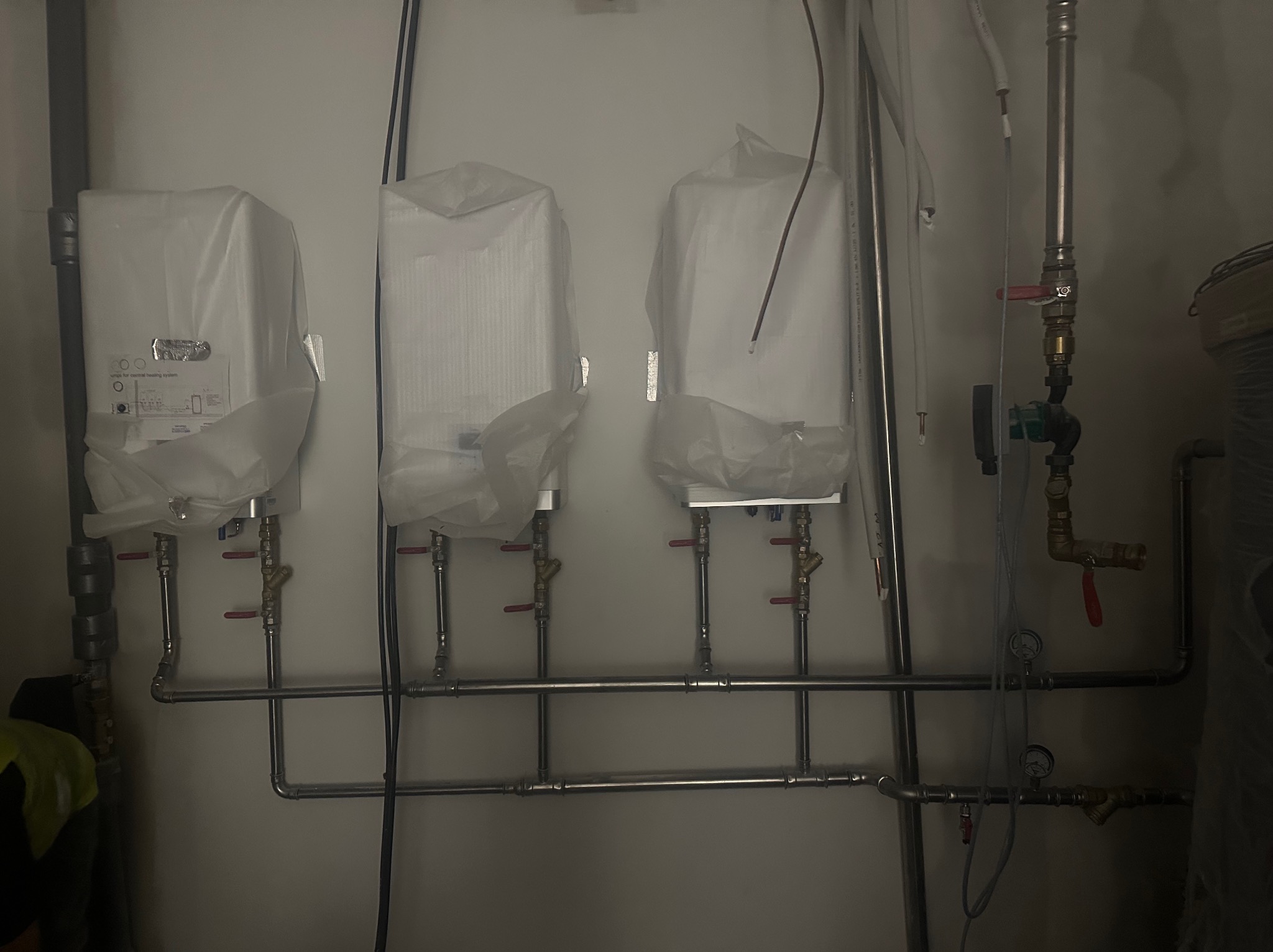 Trzy nowe grzejniki, jeszcze w folii ochronnej, zamontowane na ścianie z widocznymi rurami instalacji centralnego ogrzewania.