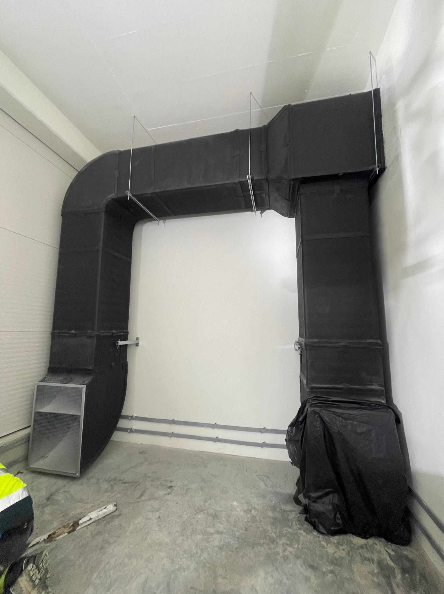 Czarny system kanałów wentylacyjnych, wsparty na metalowych linkach, zamontowany w jasnym pomieszczeniu z betonową podłogą, fragment pracownika w odblaskowej kamizelce w lewym dolnym rogu.