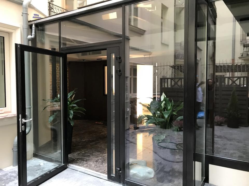 Szklana konstrukcja pawilonu z czarnymi ramami, widok na wewnętrzny dziedziniec z roślinami doniczkowymi i odbiciem osoby w szybie.