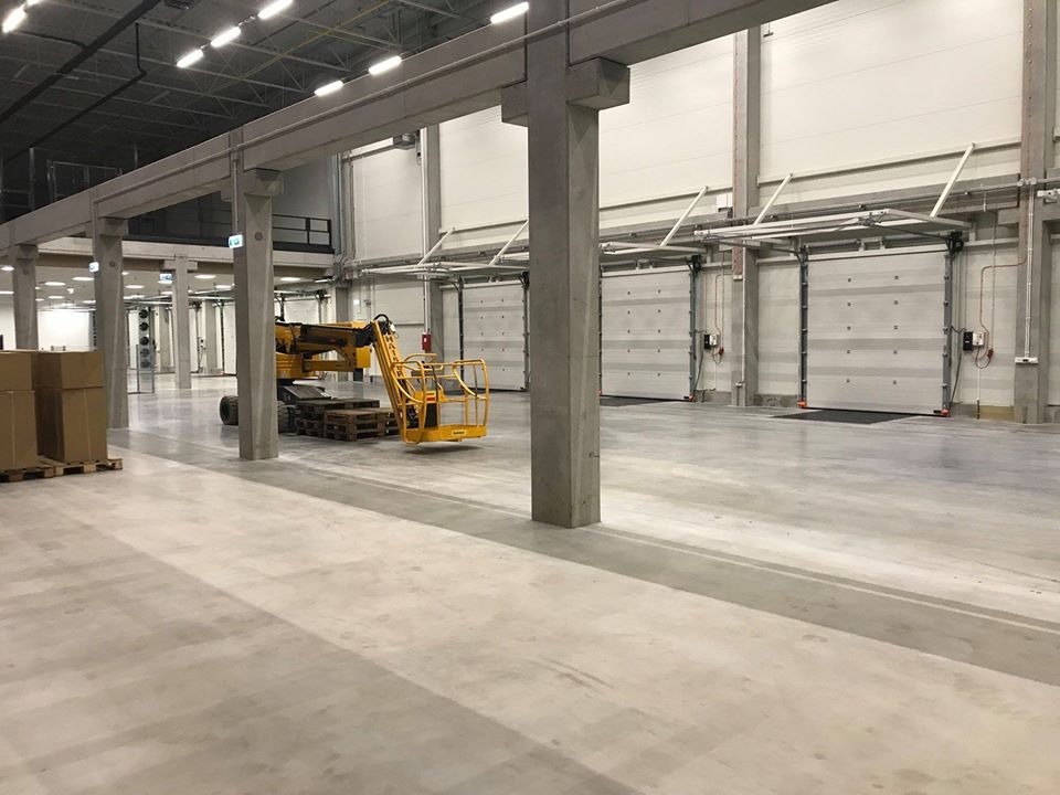 Wnętrze hali magazynowej z betonową podłogą, kolumnami i bramami segmentowymi, z żółtym podnośnikiem koszowym na paletach w tle.