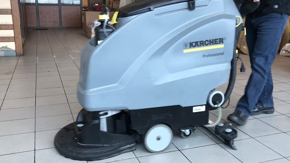 Szara, profesjonalna maszyna czyszcząca Kärcher w trakcie pracy na jasnej, kafelkowej podłodze, widoczne nogi operatora w jeansach i czarnych butach.