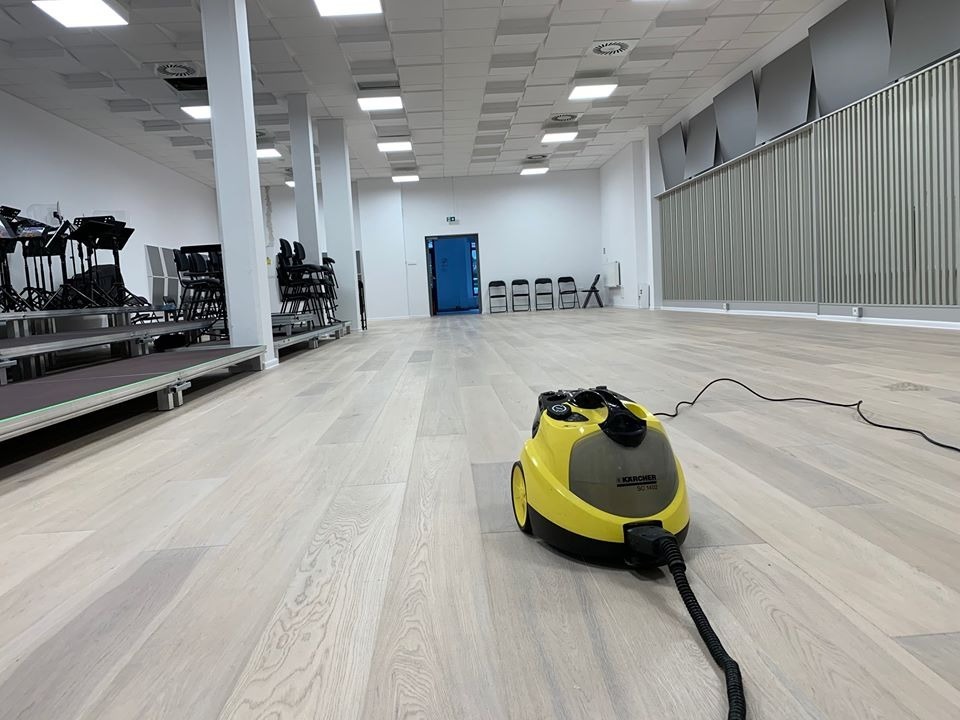 Żółty parowy mop Karcher SC 1402 na jasnej drewnianej podłodze dużej sali z białymi kolumnami, sceną i krzesłami w tle, po czyszczeniu.