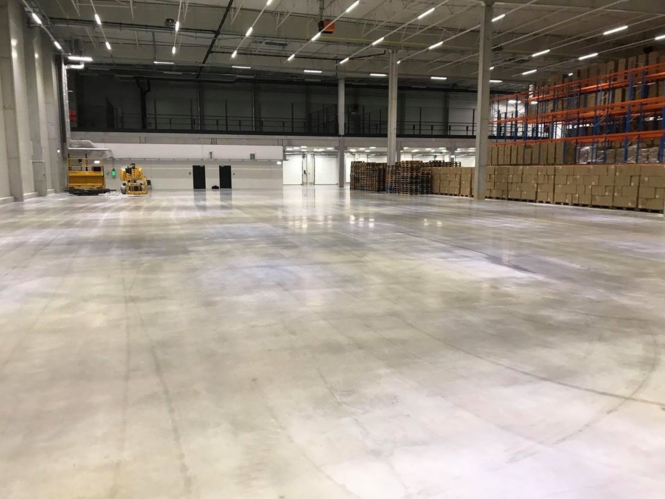 Wnętrze hali magazynowej z betonową posadzką, widoczne regały z kartonami i paletami, oraz podnośniki koszowe w oddali.