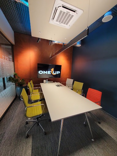 Nowoczesna sala konferencyjna z długim, białym stołem, kolorowymi krzesłami i ścianą z logo 'ONE UP'