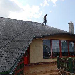 Ekspert czystości - Pracownik w kombinezonie czyści ciemny dach z bitumicznej dachówki za pomocą myjki ciśnieniowej. Widoczne efekty czyszczenia i lina zabezpieczająca.