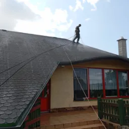 Pracownik w kombinezonie czyści ciemny dach z bitumicznej dachówki za pomocą myjki ciśnieniowej. Widoczne efekty czyszczenia i lina zabezpieczająca.