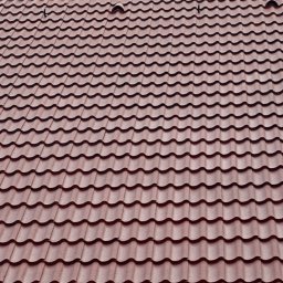 Ekspert czystości - Czerwono-brązowa dachówka ceramiczna ułożona na dachu pod kątem, widoczne elementy mocujące na szczycie. Fotografia z bliska, oświetlenie dzienne.