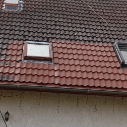 Ekspert czystości - Częściowo wyczyszczony dach z czerwonej dachówki, widoczne okna dachowe i komin. Kontrast między czystą a zabrudzoną powierzchnią. Fragment elewacji z rynną i lampą.