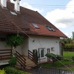 Dom jednorodzinny z brązową dachówką, białymi kominami i oknami dachowymi. Widoczna elewacja z numerem 96 i elementami zieleni. Ujęcie z poziomu gruntu.