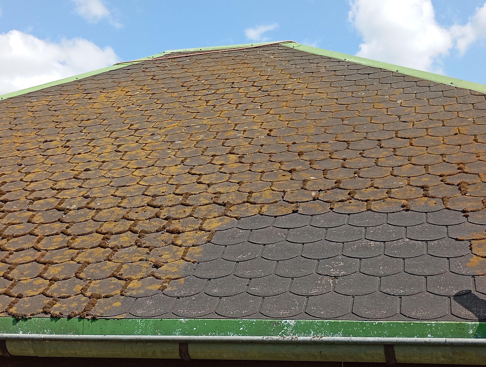 Zarośnięty mchem dach z dachówki bitumicznej, widoczne zanieczyszczenia. Ujęcie z dołu, fragment rynny. Błękitne niebo z chmurami w tle.