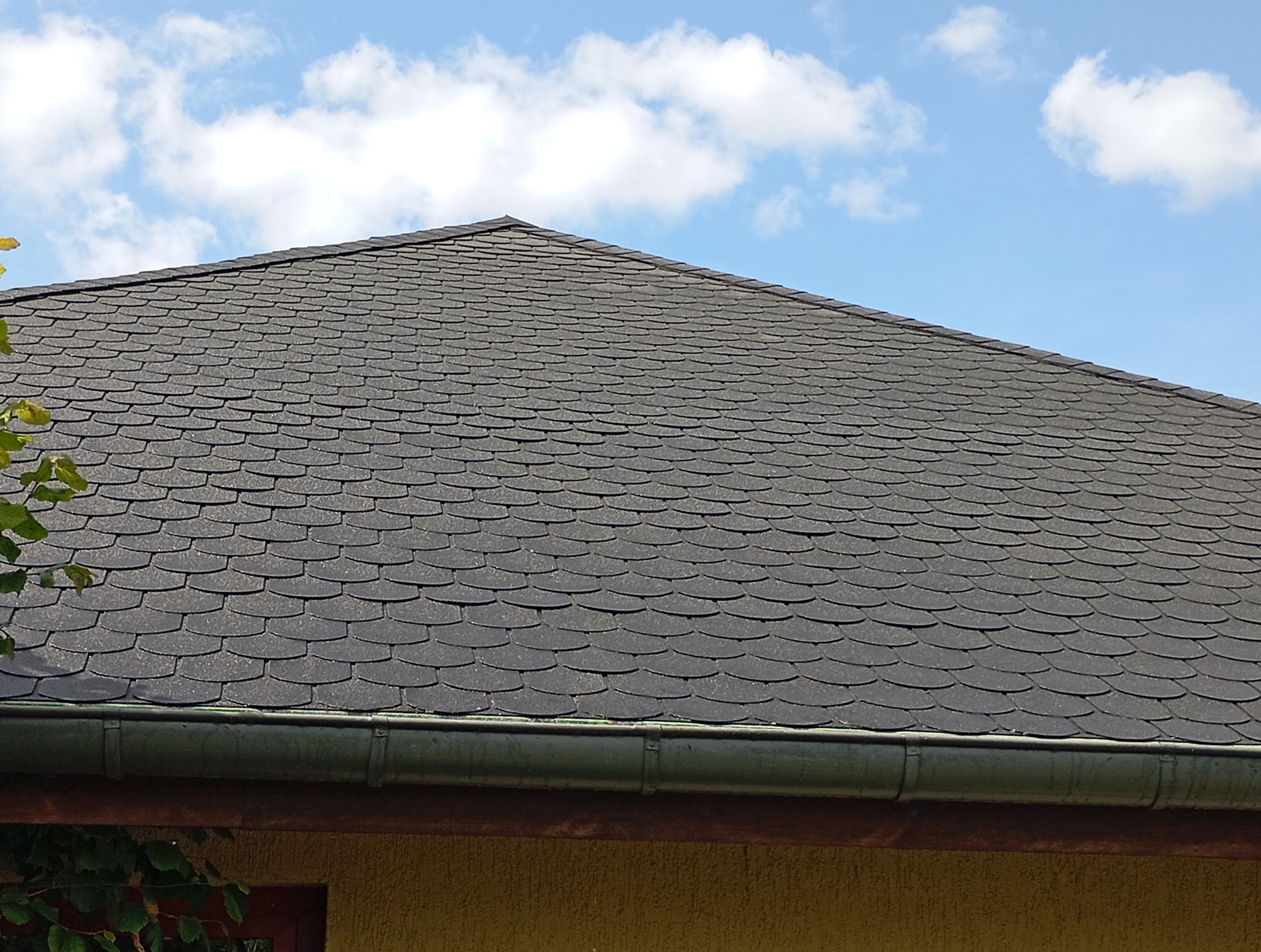 Dach pokryty czarnym gontem bitumicznym o wzorze rybiej łuski, widoczny fragment rynny i błękitne niebo z chmurami w tle. Ujęcie z dołu.