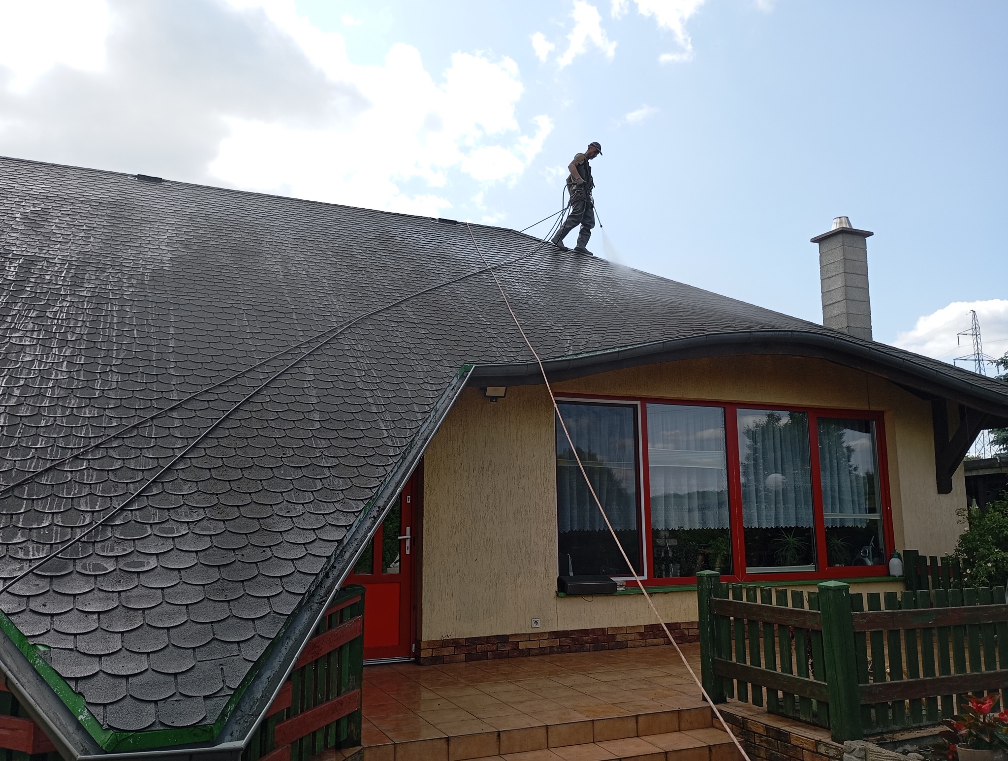 Pracownik w kombinezonie czyści ciemny dach z bitumicznej dachówki za pomocą myjki ciśnieniowej. Widoczne efekty czyszczenia i lina zabezpieczająca.