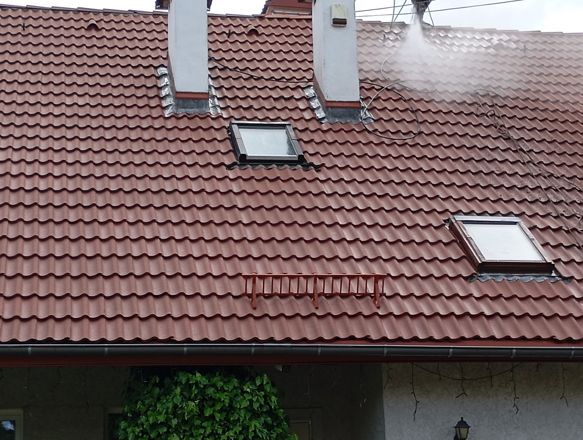 Czerwony dach z dachówką ceramiczną, czyszczony strumieniem wody pod ciśnieniem. Widoczne okna dachowe, kominy i drabinka dachowa.