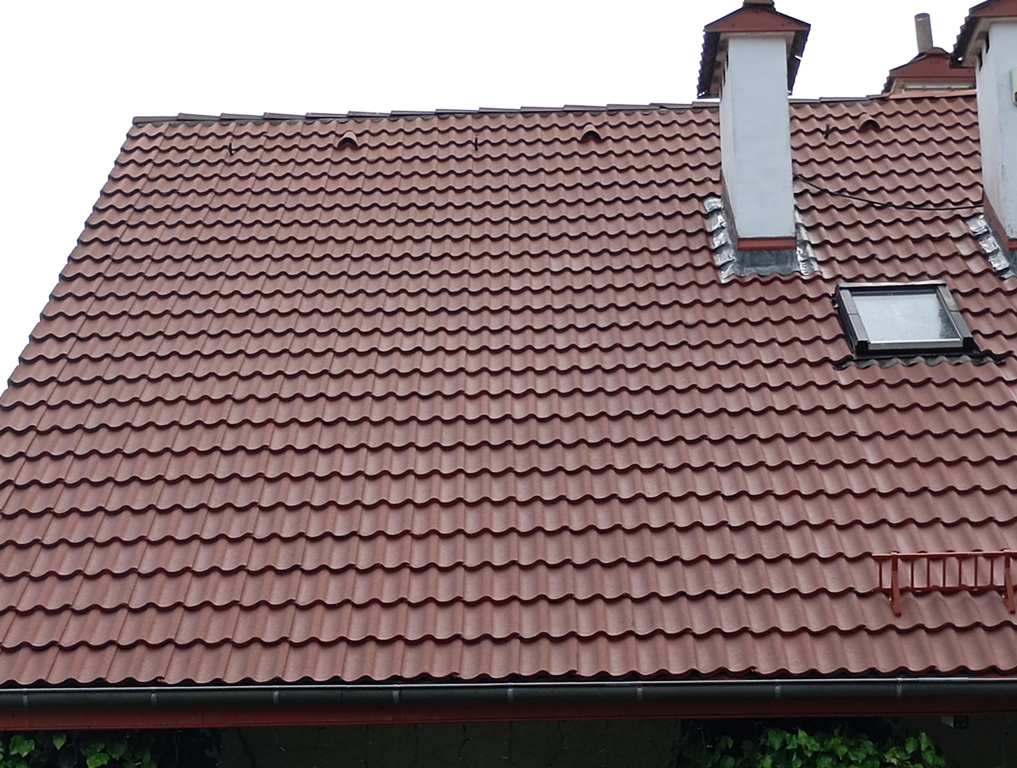 Czerwony dach z dachówki ceramicznej, widoczne kominy i okno dachowe. Ujęcie z dołu, fragment rynny i roślinności w dolnej części kadru.