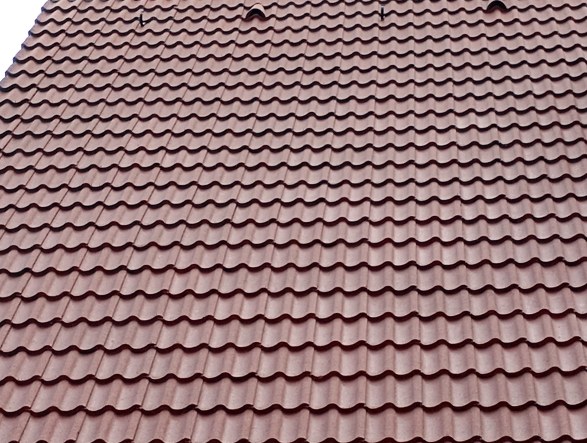 Czerwono-brązowa dachówka ceramiczna ułożona na dachu pod kątem, widoczne elementy mocujące na szczycie. Fotografia z bliska, oświetlenie dzienne.