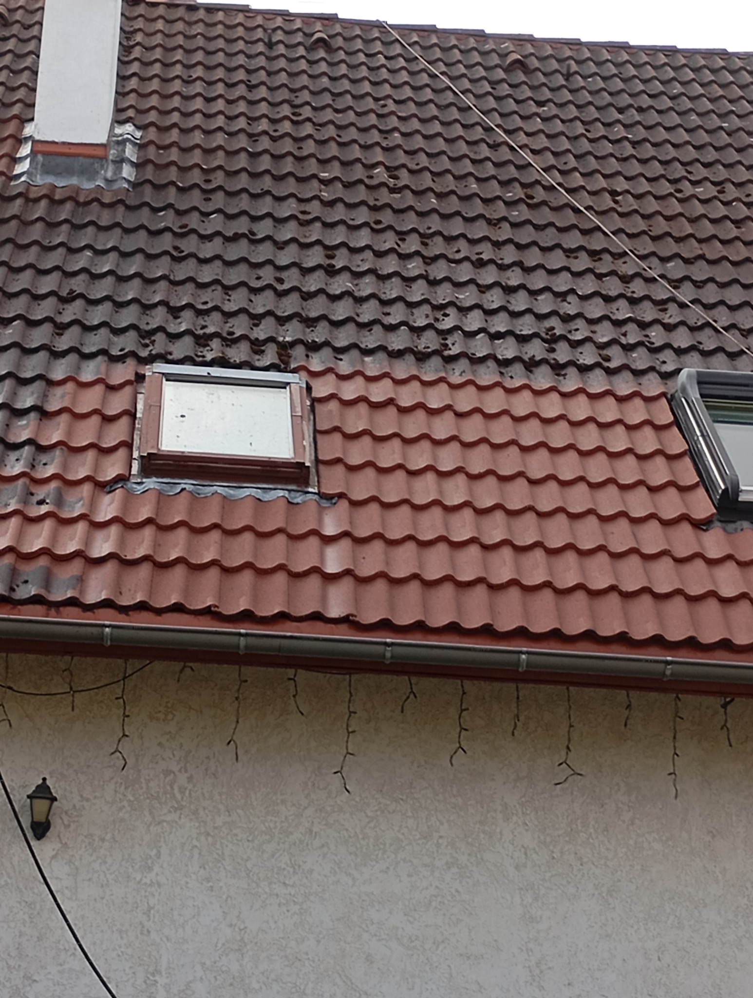 Częściowo wyczyszczony dach z czerwonej dachówki, widoczne okna dachowe i komin. Kontrast między czystą a zabrudzoną powierzchnią. Fragment elewacji z rynną i lampą.