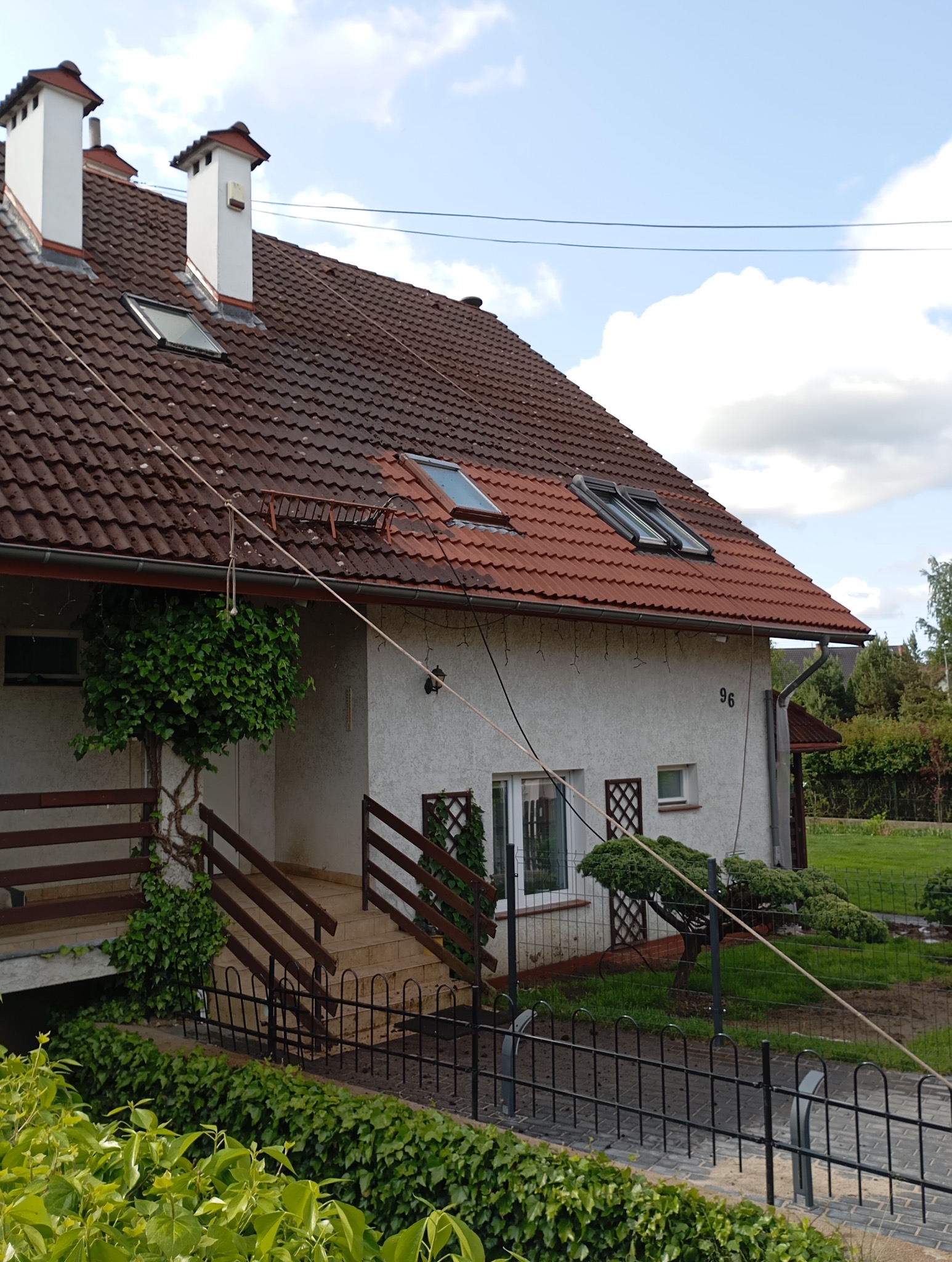 Dom jednorodzinny z brązową dachówką, białymi kominami i oknami dachowymi. Widoczna elewacja z numerem 96 i elementami zieleni. Ujęcie z poziomu gruntu.