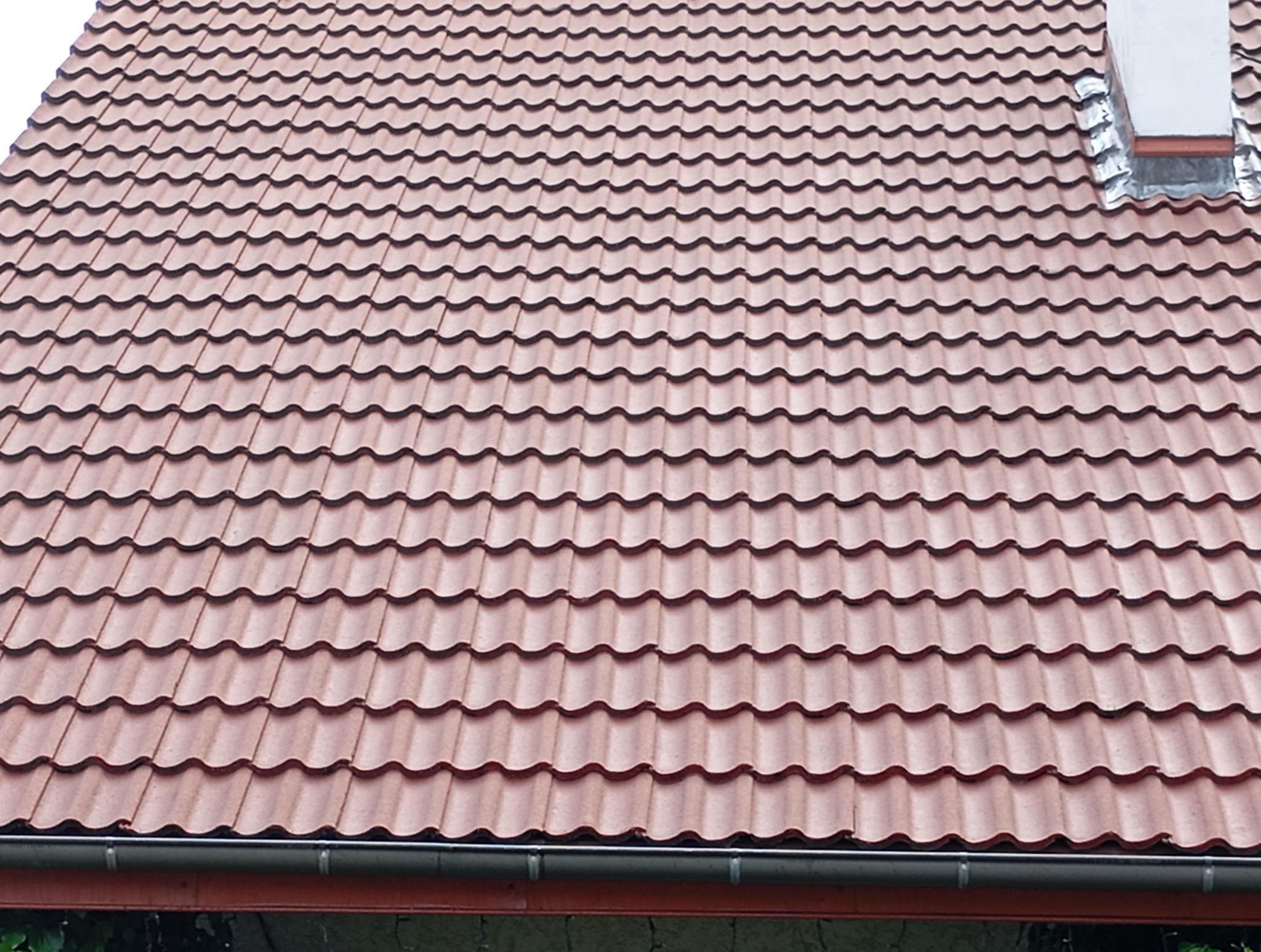 Czerwona dachówka ceramiczna na skośnym dachu domu, widok z dołu na rynnę i komin. Regularny wzór dachówek, równomierne oświetlenie.