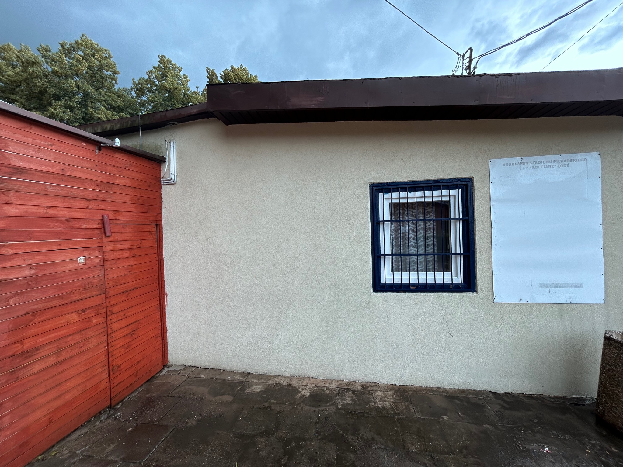 Fragment budynku z beżową elewacją, oknem z kratą i tablicą informacyjną. Widoczny fragment dachu, czerwona drewniana ściana i mokra kostka brukowa. Pochmurne niebo.
