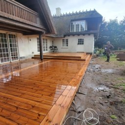 ANDBUD - Świeżo zaimpregnowany, lśniący drewniany taras przy domu z krytym strzechą dachem w Górowie Iławeckim. W tle widoczne materiały budowlane i pracownik.