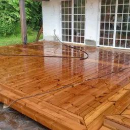 Świeżo zaimpregnowany, mokry drewniany taras z widocznymi śladami wody po myciu, z elementami dekoracyjnymi na obrzeżach, na tle białego domu z dwuskrzydłowymi oknami.