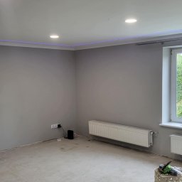 ANDBUD - Szare, świeżo pomalowane wnętrze pokoju z grzejnikami, oświetleniem sufitowym i listwą LED pod sufitem. Podłoga w trakcie przygotowania pod wykończenie, widoczny plastikowy pojemnik z narzędziami.