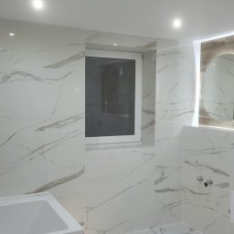 ANDBUD - Nowoczesna łazienka wykończona białymi płytkami imitującymi marmur, z widoczną umywalką z drewnopodobną szafką, oknem i okrągłym lustrem z podświetleniem.