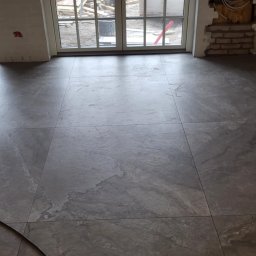 ANDBUD - Szare, wielkoformatowe płytki podłogowe imitujące kamień, ułożone w pomieszczeniu z dużym oknem balkonowym i fragmentem niezabezpieczonej instalacji elektrycznej w ścianie.